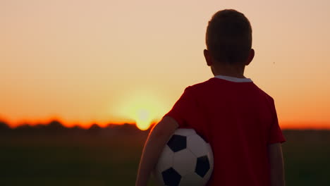 The-boy-is-on-the-field-at-sunset-in-the-grass-with-a-soccer-ball.-Dreaming-of-a-football-career.-The-concept-of-success-in-the-sport.